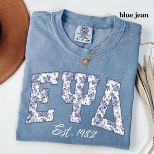 May include: A blue jean colored t-shirt with the letters "EΨA" in a floral pattern. The text "Est. 1982" is below the letters. A gold necklace with a pendant is on the shirt.