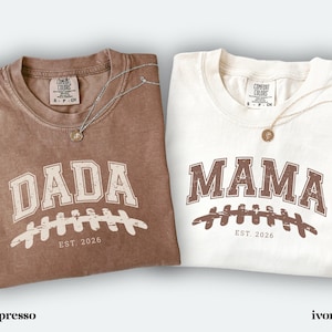 May include: Two short-sleeve crewneck t-shirts, one in espresso brown and one in ivory. The espresso shirt features the text DADA in white with a football graphic and EST. 2026. The ivory shirt features MAMA in brown with a football graphic and EST. 2026.