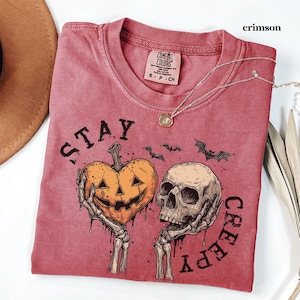 May include: A crimson t-shirt featuring a graphic print of two skeletal hands holding a heart-shaped jack-o'-lantern and a human skull. The text "STAY CREEPY" is printed in black around the illustration, which also includes small flying bats.