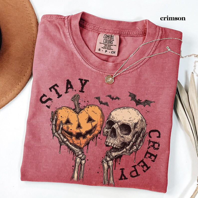 May include: A crimson t-shirt featuring a graphic print of two skeletal hands holding a heart-shaped jack-o'-lantern and a human skull. The text "STAY CREEPY" is printed in black around the illustration, which also includes small flying bats.