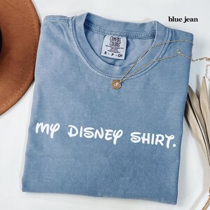 May include: A blue jean Comfort Colors t-shirt with the text "my DISNEY SHIRT." A gold necklace with a pendant rests on the shirt. The shirt is folded and has a tag on the collar.