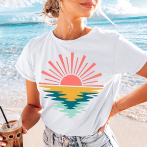 May include: White t-shirt featuring a colorful graphic of a sunset over the ocean. The design includes a pink sun with orange rays, a yellow horizon line, and blue and green water reflections. The shirt is made of a soft material.