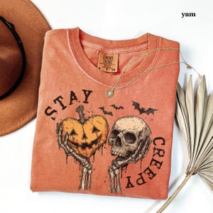 May include: A burnt orange t-shirt featuring a graphic print of two skeletal hands holding a heart-shaped jack-o'-lantern and a human skull. The text "STAY CREEPY" is printed in a dark font around the graphic, accompanied by small bat silhouettes.