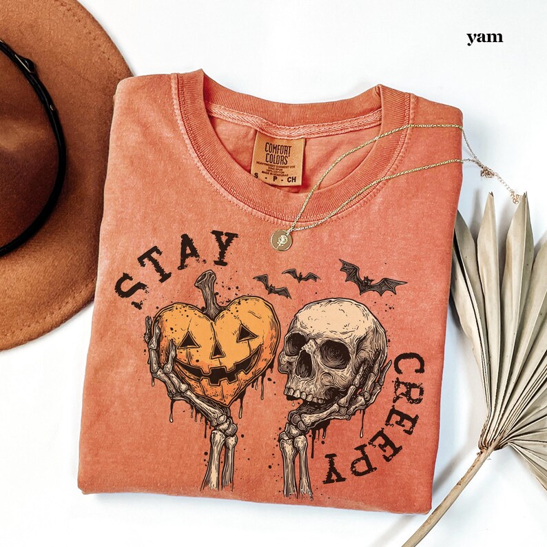 May include: A burnt orange t-shirt featuring a graphic print of two skeletal hands holding a heart-shaped jack-o'-lantern and a human skull. The text "STAY CREEPY" is printed in a dark font around the graphic, accompanied by small bat silhouettes.