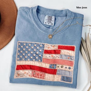 May include: Pale blue t-shirt with a patchwork American flag design. The flag is made of red, white, and blue fabric patches with stars and stripes. The shirt has a crew neck and short sleeves. A gold necklace with a pendant is on the shirt.