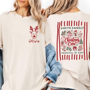 May include: A cream-colored t-shirt featuring a red reindeer graphic and the name Olivia on the front. The back displays a red-striped border containing Christmas icons, the text Smith Family, Christmas Crew, and Charleston, SC 2026.