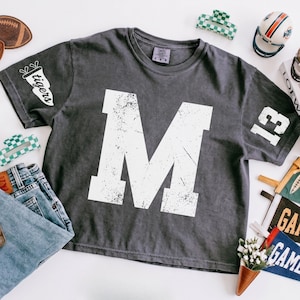 May include: A charcoal gray short-sleeve t-shirt featuring a large, distressed white letter M on the front. The right sleeve displays the word tigers, while the left sleeve features the number 13 in white, distressed block lettering.