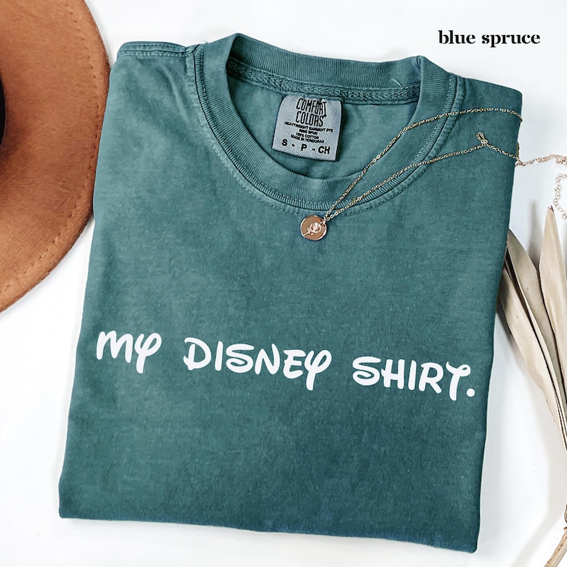 May include: A blue spruce Comfort Colors t-shirt with the text "my DISNEY SHIRT" in white. A gold necklace with a pendant is draped over the shirt. The shirt is folded and placed on a white surface.