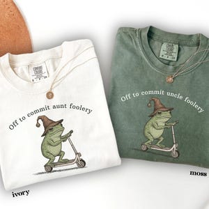 May include: Two t-shirts, ivory and moss green, feature a frog riding a scooter and wearing a wizard hat. The text on the ivory shirt reads "Off to commit aunt foolery," and the moss shirt says "Off to commit uncle foolery."