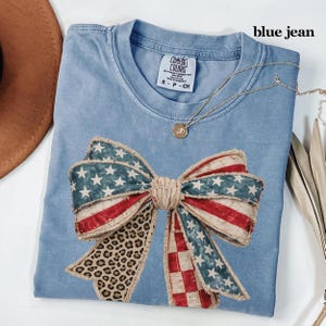 May include: A light blue t-shirt with a large bow design featuring American flag and leopard print patterns. The shirt has the text "blue jean" and a gold necklace. The shirt is made of Comfort Colors heavyweight garment dye cotton.