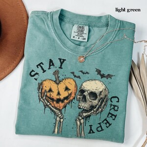 May include: A light green short-sleeved t-shirt with a graphic print featuring two skeletal hands holding a heart-shaped jack-o'-lantern and a human skull. The text "STAY CREEPY" is printed around the graphic, which also includes small bats.