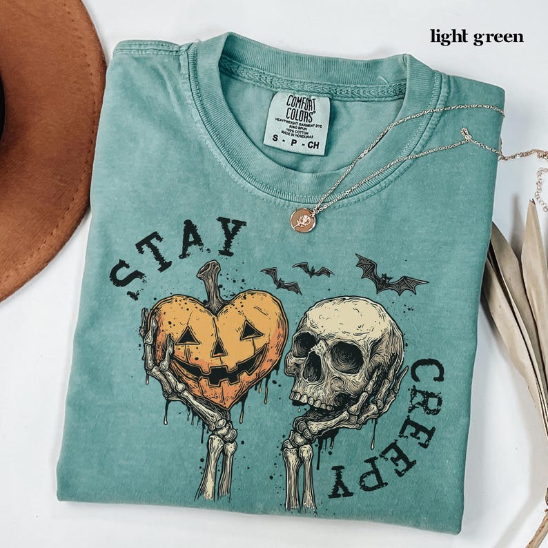 May include: A light green short-sleeved t-shirt with a graphic print featuring two skeletal hands holding a heart-shaped jack-o'-lantern and a human skull. The text "STAY CREEPY" is printed around the graphic, which also includes small bats.