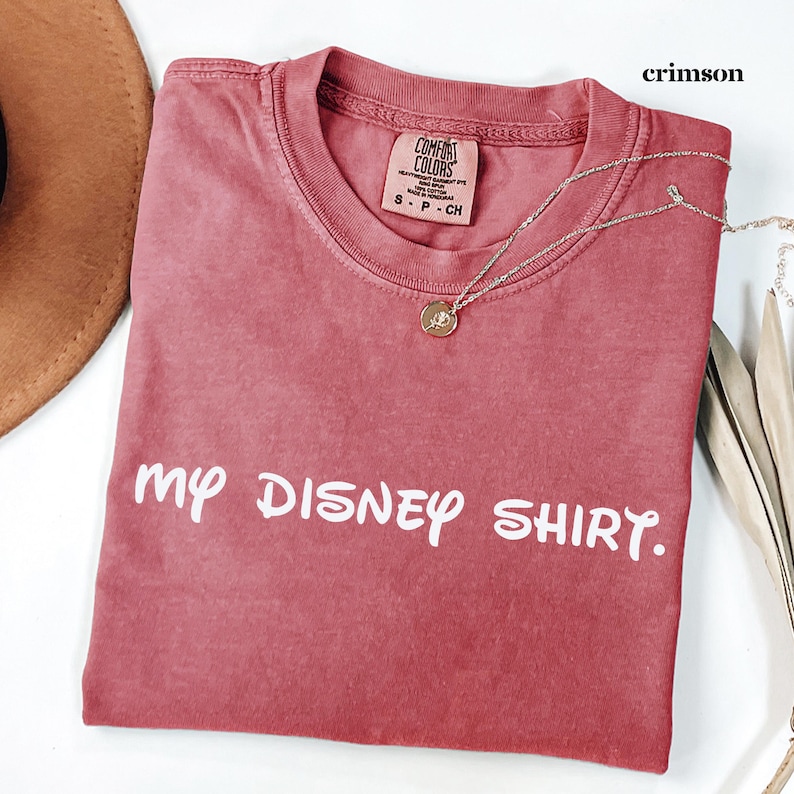 May include: Crimson-colored t-shirt with the text "my DISNEY SHIRT." A silver necklace with a round pendant is draped over the shirt. The shirt has a crew neck and a tag that reads "COMFORT COLORS."
