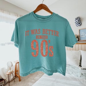 May include: A teal short-sleeved crew neck t-shirt featuring the text "IT WAS BETTER IN THE 90's" printed in a distressed, faded coral-orange color across the chest. The shirt is displayed on a wooden hanger against a neutral-colored bedroom background.