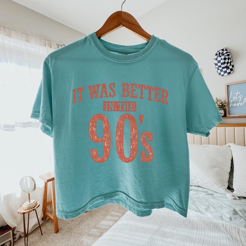 May include: A teal short-sleeved crew neck t-shirt featuring the text "IT WAS BETTER IN THE 90's" printed in a distressed, faded coral-orange color across the chest. The shirt is displayed on a wooden hanger against a neutral-colored bedroom background.