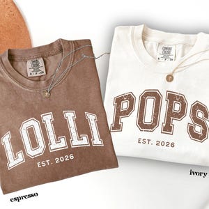 May include: Two t-shirts, one espresso and one ivory, with the words "LOLLI" and "POPS" printed in a collegiate style. Both shirts have "EST. 2026" printed below the main text. Each shirt has a silver necklace with a pendant.