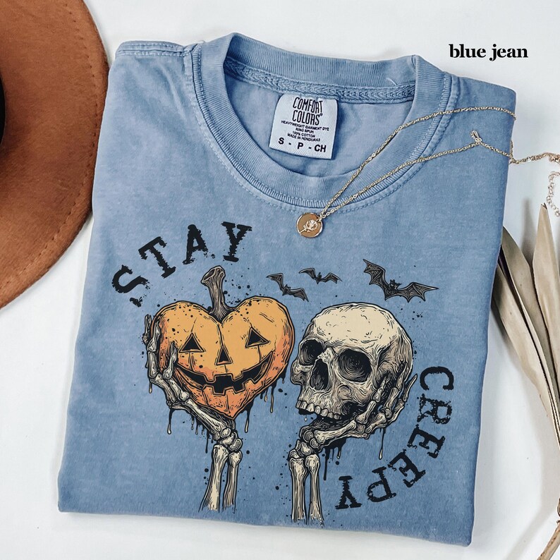 May include: A blue jean coloured t-shirt featuring a graphic of two skeletal hands holding a heart-shaped pumpkin and a human skull. The text "STAY CREEPY" is printed around the illustration, accompanied by small bat silhouettes and a gold necklace.