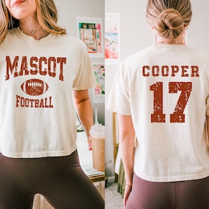 May include: A cream-colored short-sleeve t-shirt featuring a distressed red graphic on the front that reads MASCOT FOOTBALL with a football icon. The back of the shirt displays the name COOPER and the number 17 in the same distressed red font.