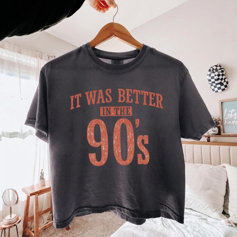 May include: A charcoal gray short-sleeve t-shirt featuring the orange distressed text "IT WAS BETTER IN THE 90'S" printed on the front. The shirt has a crew neckline and a relaxed fit, displayed on a wooden hanger against a neutral background.