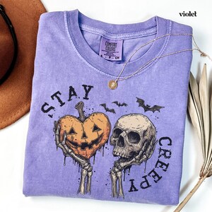 May include: A violet short-sleeve t-shirt featuring a graphic print of two skeletal hands holding a heart-shaped jack-o'-lantern and a human skull. The text "STAY CREEPY" is printed in black around the graphic, accompanied by small bat silhouettes.