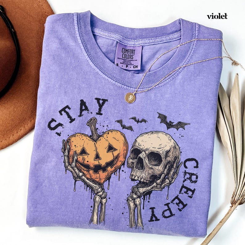May include: A violet short-sleeve t-shirt featuring a graphic print of two skeletal hands holding a heart-shaped jack-o'-lantern and a human skull. The text "STAY CREEPY" is printed in black around the graphic, accompanied by small bat silhouettes.