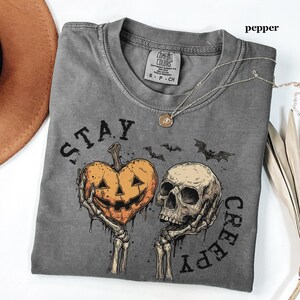 May include: A pepper-coloured short-sleeve t-shirt featuring a graphic print of two skeletal hands holding a heart-shaped jack-o'-lantern and a human skull. The text "STAY CREEPY" is printed in black around the graphic, which also includes two small bats.