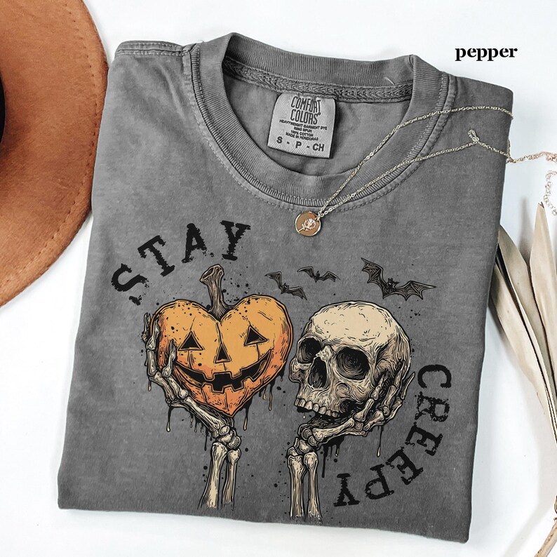 May include: A pepper-coloured short-sleeve t-shirt featuring a graphic print of two skeletal hands holding a heart-shaped jack-o'-lantern and a human skull. The text "STAY CREEPY" is printed in black around the graphic, which also includes two small bats.