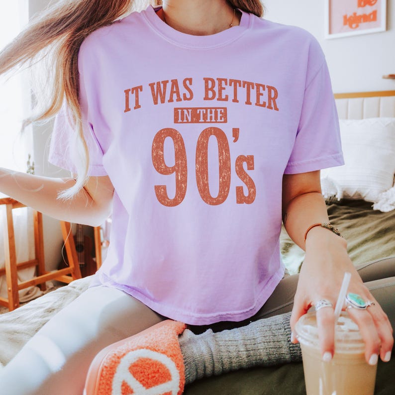 May include: A light purple short-sleeve t-shirt featuring the text "IT WAS BETTER IN THE 90's" printed in a distressed, burnt orange font across the chest. The shirt has a crew neckline and a relaxed fit, showcasing a vintage-inspired graphic design.