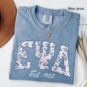 May include: Blue jean Comfort Colors t-shirt with floral design letters spelling out EΨA. A pink bow is centered below the letters. The text "Est. 1982" is below the letters. A gold necklace with a pendant is on the shirt.