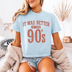 May include: A light blue short-sleeve t-shirt featuring the text "IT WAS BETTER IN THE 90's" printed in a distressed, reddish-brown font across the chest. The shirt has a crew neckline and a relaxed fit, showcasing a vintage-inspired graphic design.