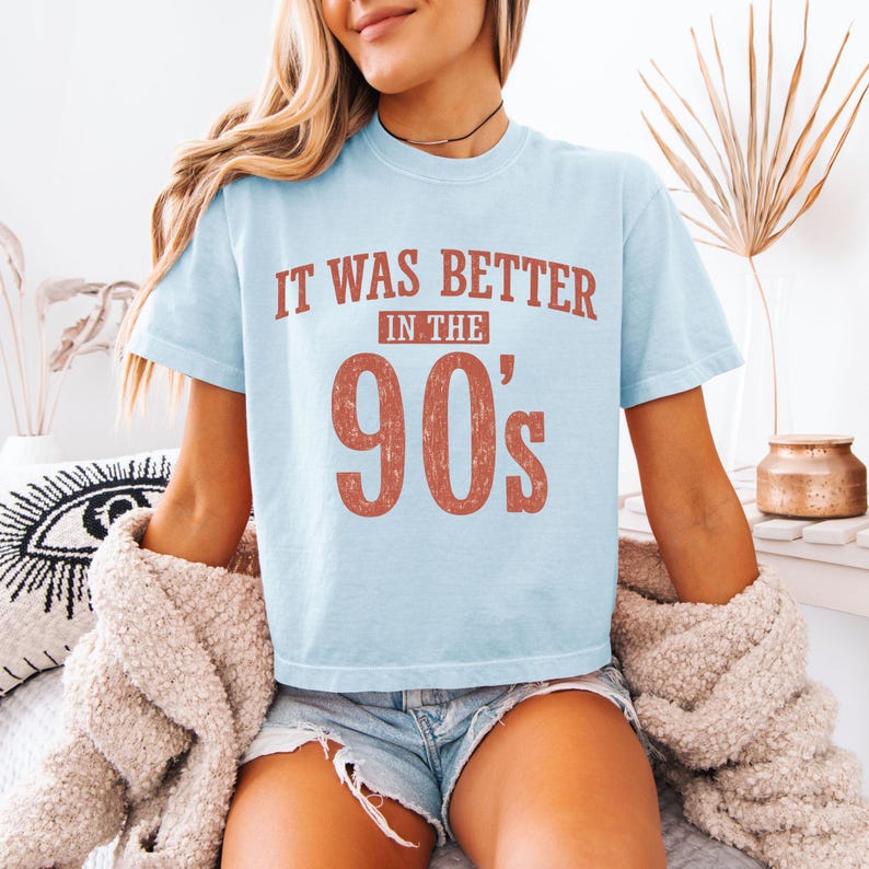 May include: A light blue short-sleeve t-shirt featuring the text "IT WAS BETTER IN THE 90's" printed in a distressed, reddish-brown font across the chest. The shirt has a crew neckline and a relaxed fit, showcasing a vintage-inspired graphic design.