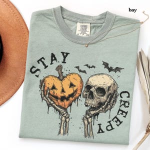 May include: A sage green t-shirt featuring a graphic print of two skeletal hands holding a heart-shaped jack-o'-lantern and a human skull. The text "STAY CREEPY" is arched around the design, which also includes small black bats and ink splatters.