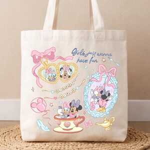 May include: A cream-colored canvas tote bag featuring a graphic print of Daisy Duck and Minnie Mouse. The design includes a heart-shaped locket, a teacup, a mirror, and the text "Girls just wanna have fun" and "DISNEY GIRLS" in a playful style.