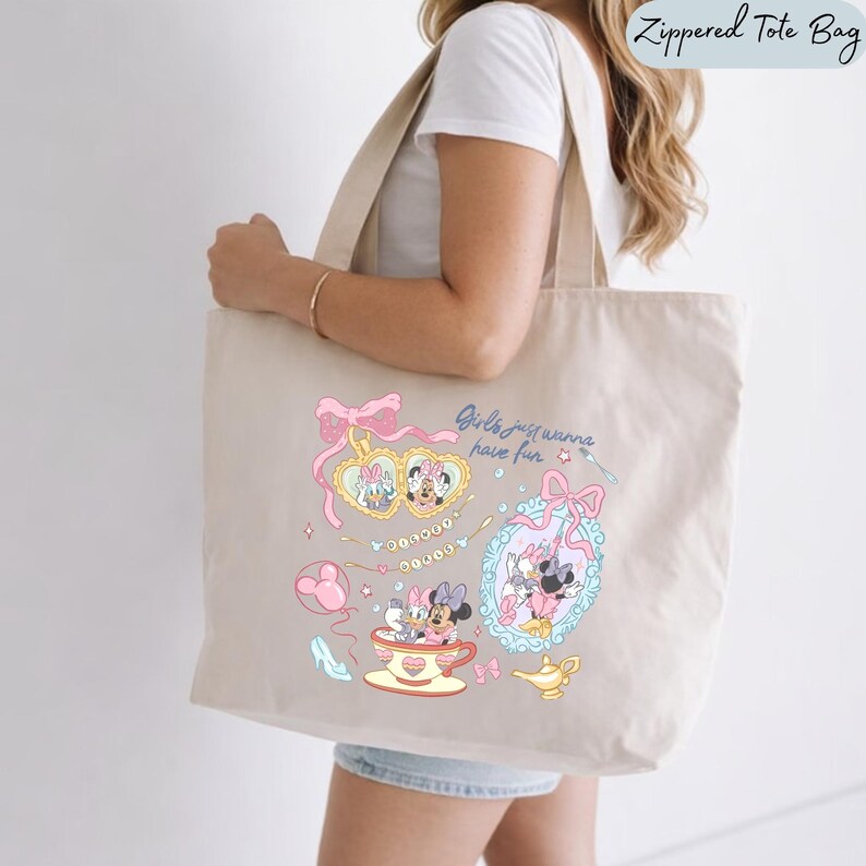 May include: A beige canvas tote bag featuring a colorful graphic print of cartoon characters in heart-shaped frames, a teacup, and a magic lamp. The design includes the text "Girls just wanna have fun" in a blue script font and pink bows.