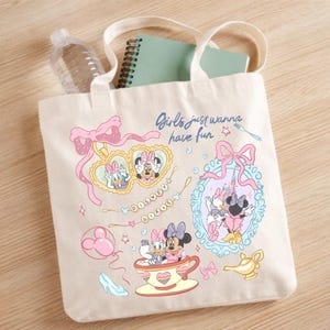 May include: A cream-colored canvas tote bag featuring a colorful graphic print of Minnie Mouse and Daisy Duck. The design includes the text 'Girls just wanna have fun', a heart-shaped locket, a teacup, a magic lamp, and various pink bows.