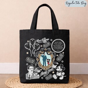 May include: A black tote bag featuring a white graphic print of Walt Disney, Mickey Mouse, balloons, park tickets, and a circular quote that reads: Here you leave today and enter the world of yesterday, tomorrow and fantasy. The design includes a central ornate gold-toned frame containing an illustration of Walt Disney holding Mickey Mouse's hand.