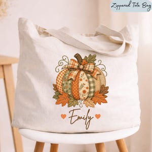 May include: A cream-colored canvas zippered tote bag featuring a central graphic of a pumpkin composed of various patterned fabric segments in orange, green, and brown. The name Emily is printed below in brown script with two orange hearts.