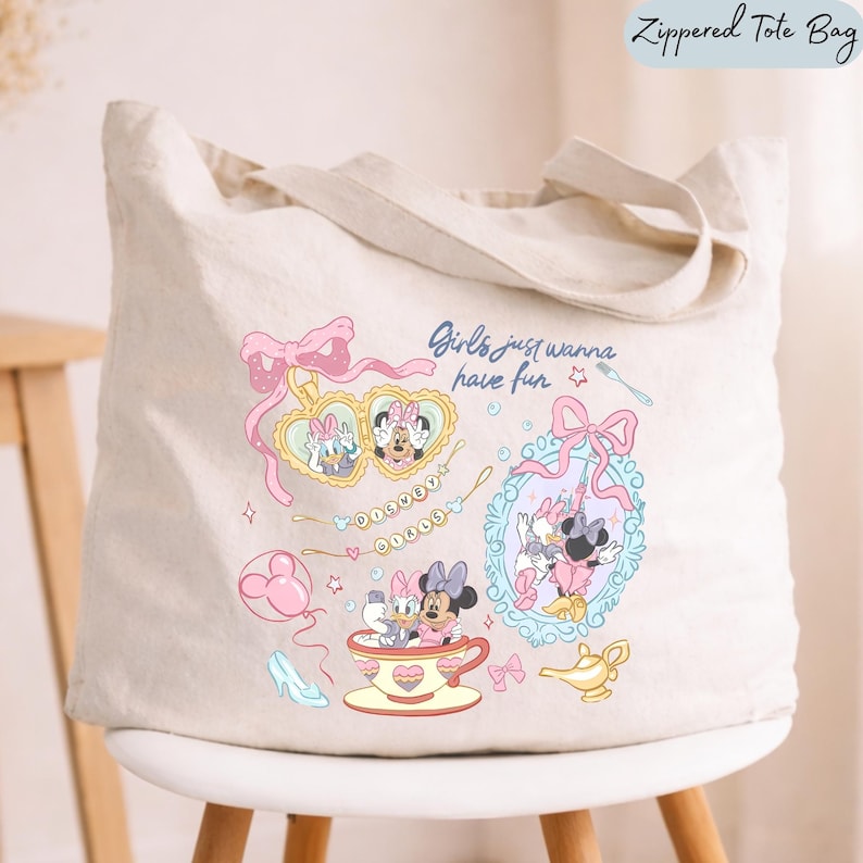 May include: A cream-colored canvas zippered tote bag featuring a colorful graphic print of Daisy Duck and Minnie Mouse. The design includes a heart-shaped locket, a teacup, a mirror, and the text "Girls just wanna have fun" and "DISNEY GIRLS".