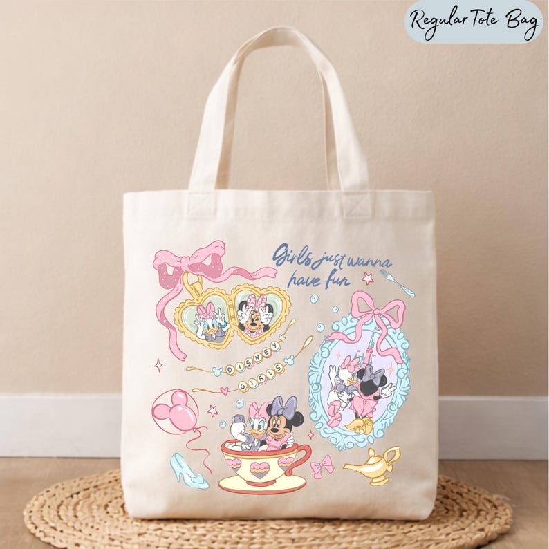 May include: A cream-colored canvas tote bag featuring a graphic print of Daisy Duck and Minnie Mouse. The design includes a heart-shaped locket, a teacup, a mirror, and the text "Girls just wanna have fun" and "DISNEY GIRLS" in a playful style.