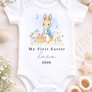 May include: White baby onesie with a watercolor illustration of a bunny wearing a blue jacket, holding a basket of Easter eggs. Text reads "My First Easter" and a name and year. The onesie is surrounded by flowers and butterflies.