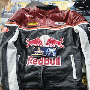 May include: A black, red, and white leather motorcycle jacket featuring the Red Bull logo, two red bull graphics, and sponsor patches including Motul, Michelin, and Helmet. A rectangular card with the text Motorbike Wears is placed on the front.
