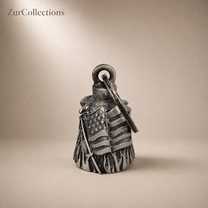 May include: A silver-toned motorcycle bell with an American flag design. The bell features a loop for hanging and a detailed depiction of the flag with stars and stripes. The base of the bell has a flame-like design.