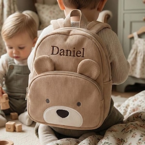 Peut inclure: Sac à dos en velours côtelé beige avec un motif de visage d'ours, brodé du nom "Daniel". Le sac à dos a des oreilles d'ours et une poche avant. L'arrière-plan montre un enfant portant le sac à dos.