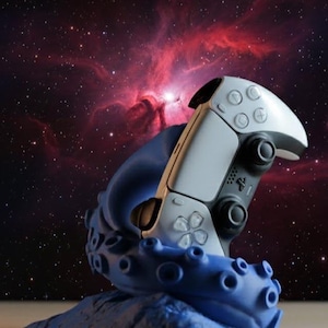 May include: A white and black video game controller rests on a blue octopus-shaped stand. The controller has multiple buttons and a central touchpad. The background features a nebula with red and purple hues, creating a space-themed aesthetic.