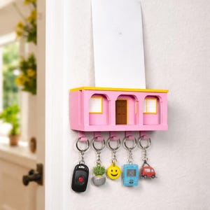 Peut inclure: Un porte-clés rose avec un toit jaune et des ouvertures en arc, ressemblant à un bâtiment miniature. Il comporte une porte marron et deux fenêtres encadrées de jaune. Cinq clés sont suspendues en dessous, chacune avec un charme unique, dont une clé de voiture, une plante, un smiley, un jeu bleu et une voiture rouge.