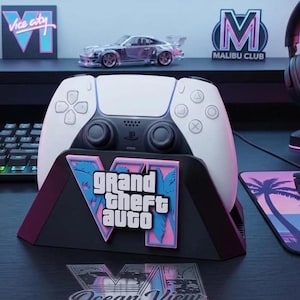 Może przedstawiać: Biało-czarny kontroler do gier w czarnej stacji ładującej z różowo-niebieskim logo Grand Theft Auto VI. Stacja znajduje się na ciemnej powierzchni, z przodu widoczna jest przezroczysta naklejka Ocean View oraz akcesoria do gier.