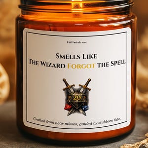 May include: A lit candle in a brown glass jar with a white label. The label reads "Smells Like The Wizard Forgot The Spell" with a graphic of crossed swords and a die. The candle is on a gray stone surface.