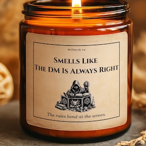Könnte beinhalten: Ein brennendes braunes Glas-Kerzenglas mit einem Etikett mit der Aufschrift "SMELLS LIKE THE DM IS ALWAYS RIGHT". Das Etikett zeigt eine Grafik einer Kapuzenfigur und Bücher. Die Kerze steht auf einer grauen Steinoberfläche.