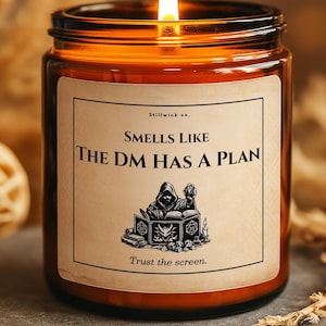 May include: A lit candle in a brown glass jar with a label that reads "Smells Like The DM Has A Plan." The label features an illustration of a hooded figure. The candle sits on a grey stone surface.