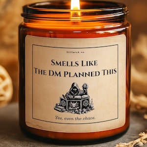 May include: A lit brown glass candle jar with a label that reads "Smells Like The DM Planned This." The label features an illustration of a cloaked figure and books. The candle is on a gray stone surface.
